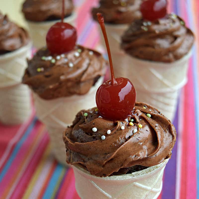 Group of ice cream cones filled with chocolate cupcake and topped with chocolate frosting, sprinkles and a cherry